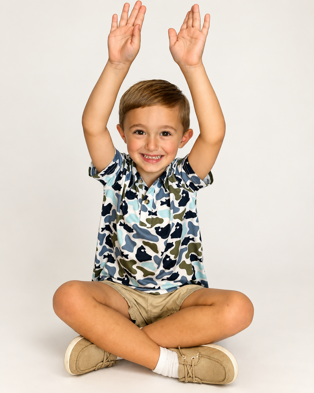 Boy sitting with arms up, wearing The Castaway Camo Polo in blue and green fish camo