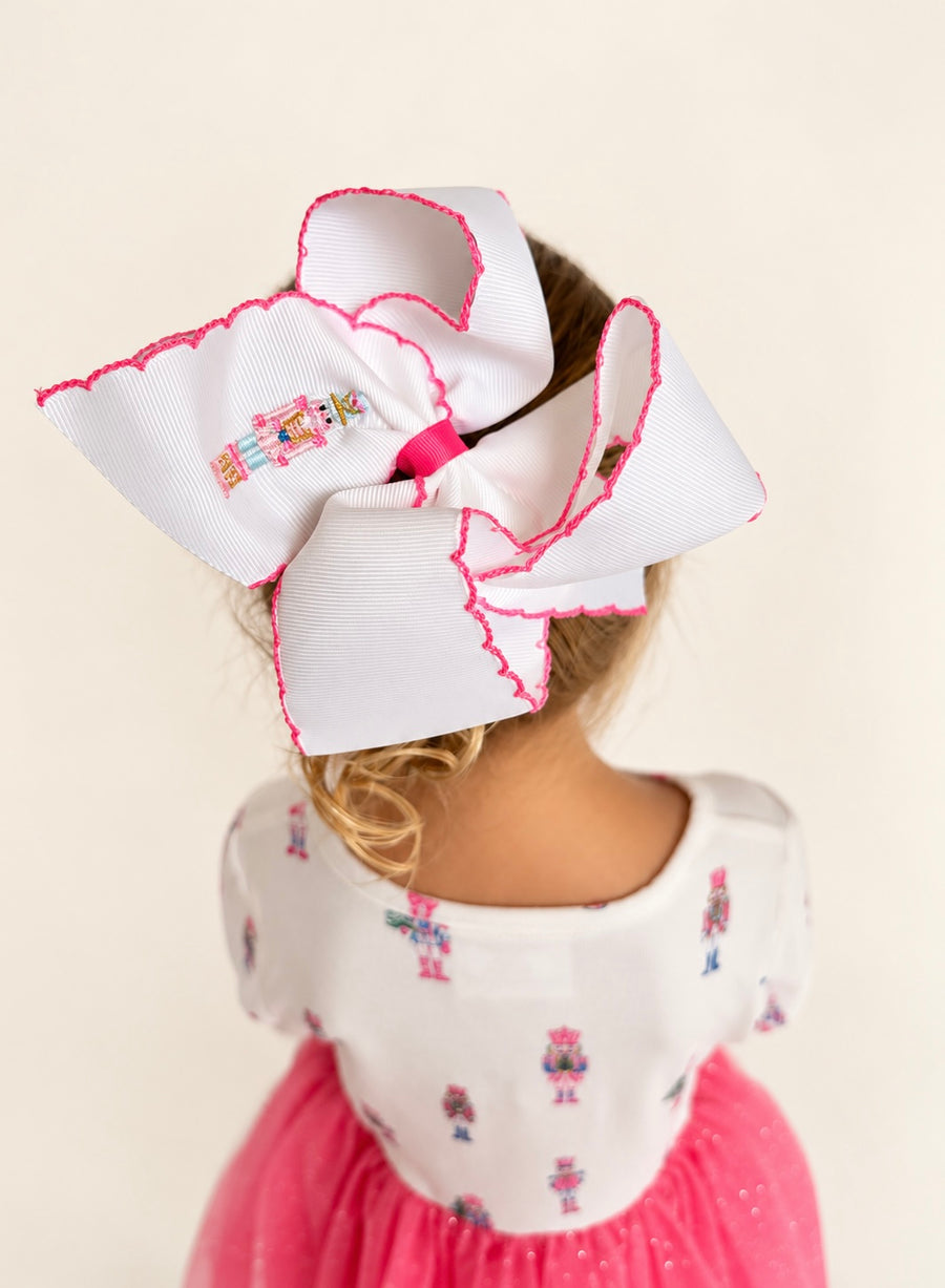 The Nutcracker Bow, white hair bow with pink scalloped trim and embroidered nutcracker, worn by a girl